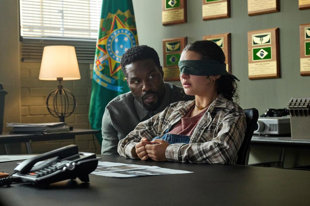 Yahya Abdul-Mateen II as John Creasy leans over Billie Boulet as Poe Rayburn seated at a table in an office setting with a Brazilian flag in the background.