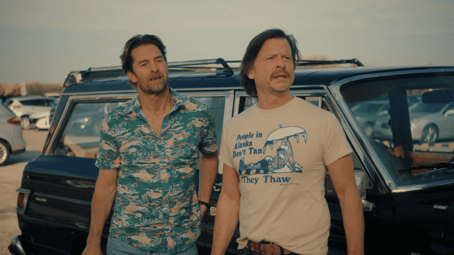 Scott Speedman as RJ Decker and Kevin Rankin as Aloysius 'Wish' Aiken stand next to a vintage station wagon in an outdoor parking lot; the man on the left wears a green floral shirt, and the man on the right wears a beige t-shirt with a graphic that reads "People in Alaska Don't Tan They Thaw."