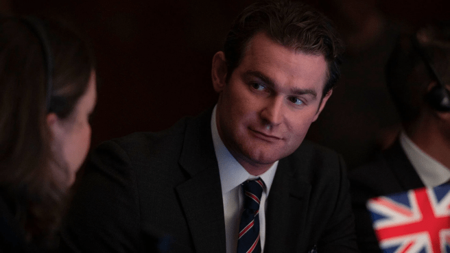 Mark Stanley in a dark suit and striped tie looks to his left, with the corner of a Union Jack flag visible on the right side of the frame. in Secret Service