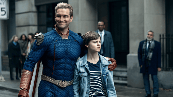 Antony Starr as Homelander in his blue superhero costume with red accents with his arm around Cameron Crovetti as Ryan wearing a denim jacket over a striped shirt, with other people blurred in the background on a city street.
