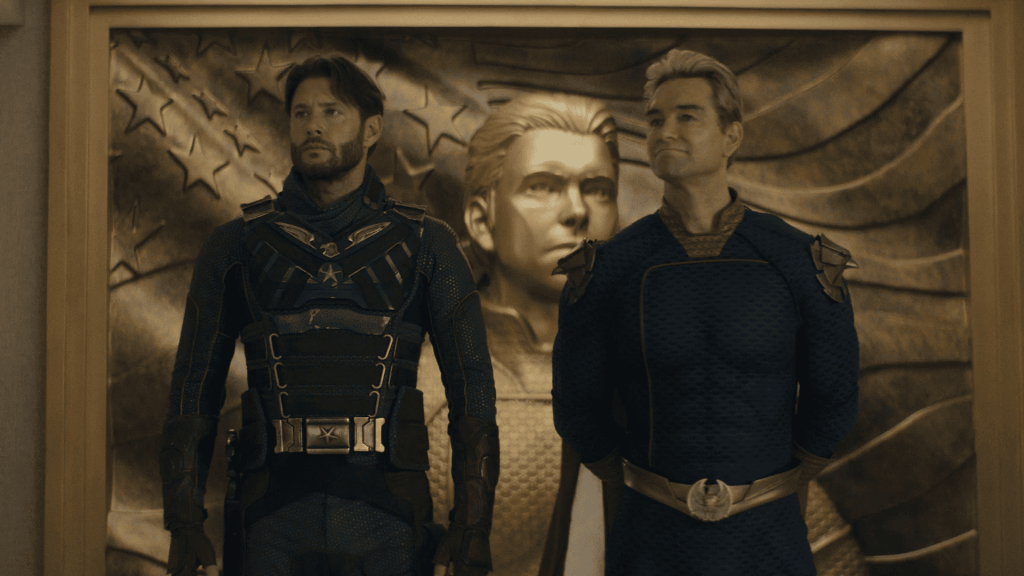 Jensen Ackles as Soldier Boy and Antony Starr as Homelander stand in front of a large golden relief sculpture of an American flag and a bust of a man. The Boys season 5