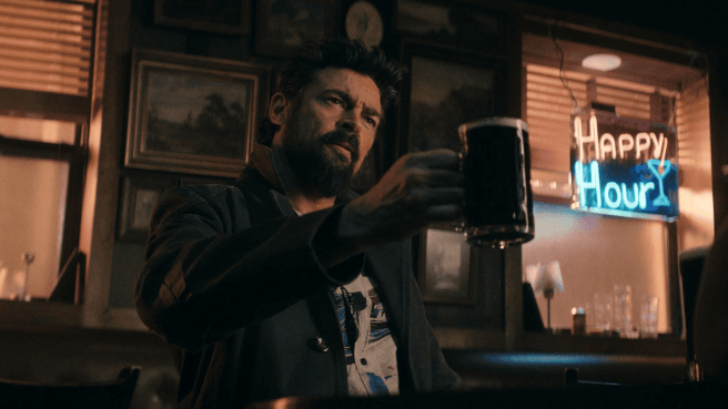 Karl Urban as Billy Butcher holding up a Guiness, toasting in a dimly lit bar with a "Happy Hour" neon sign in the background. The Boys season 5