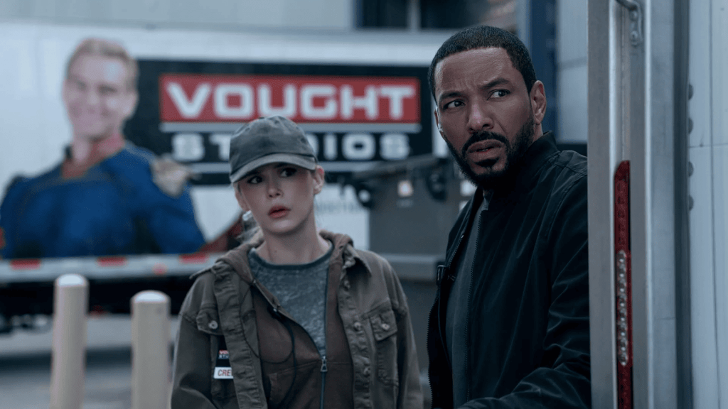Erin Moriarty as Starlight and Laz Alonso as Mother's Milk stand in front of a truck with a large image of a woman in a superhero costume and the "VOUGHT STUDIOS" logo. The Boys season 5