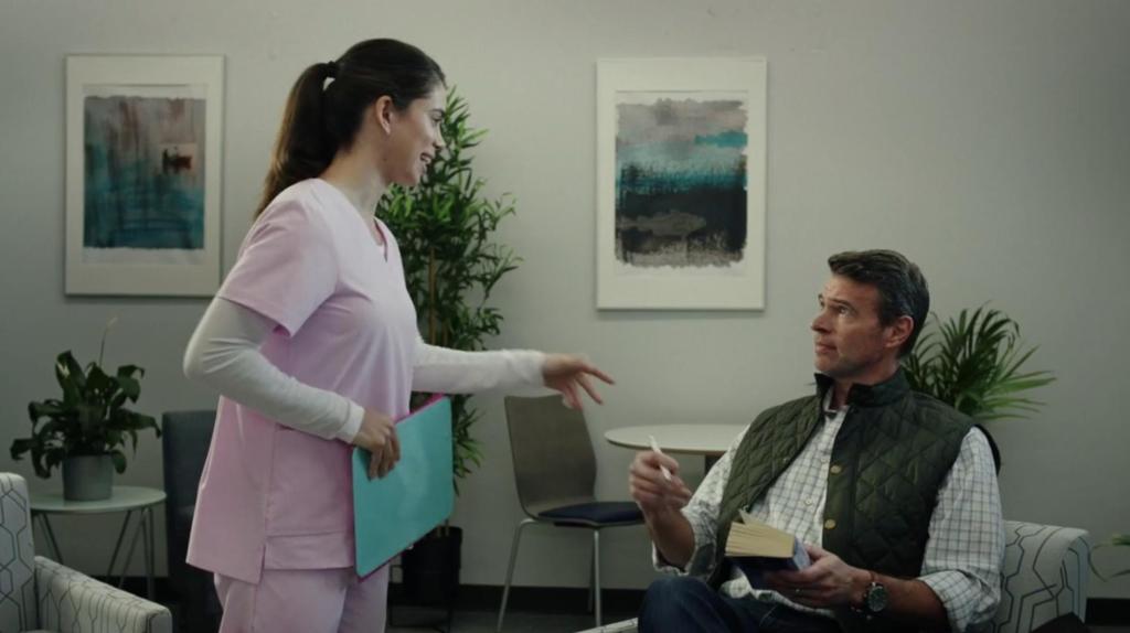 A woman in a pink medical uniform holds a teal folder and gestures to Scott Foley as Daniel wearing a green vest and plaid shirt sitting in a waiting room.