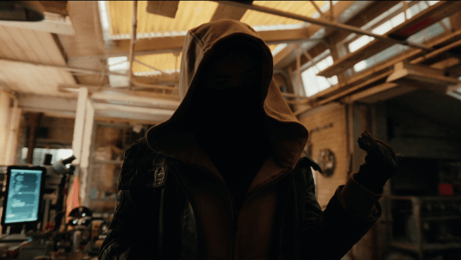 Still from SPider-Mand Brand New Day featuring, a shadowy figure wearing a hooded jacket and a dark face covering stands in a cluttered workshop, holding out a gloved hand.